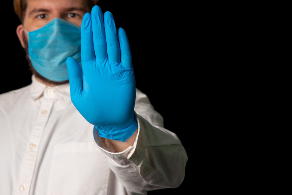 a medical professional wears a mask and holds up their hand with a blue medical glove on