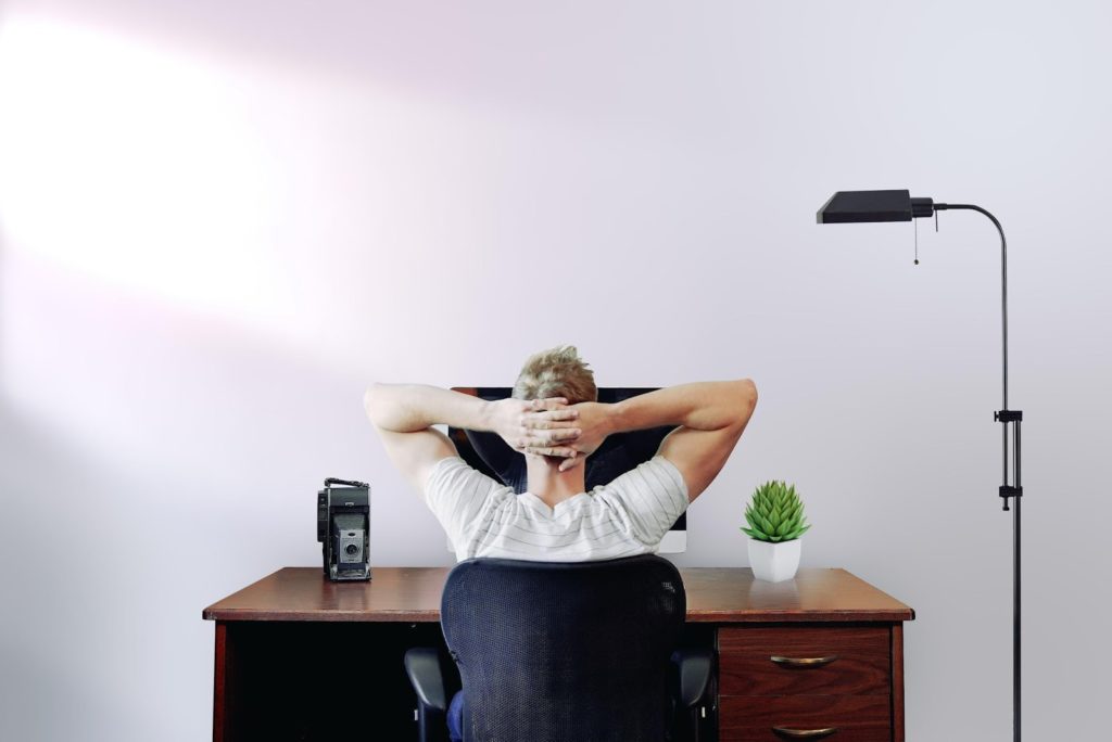 a person stares at a blank wall with their arms folded on their head at their desk