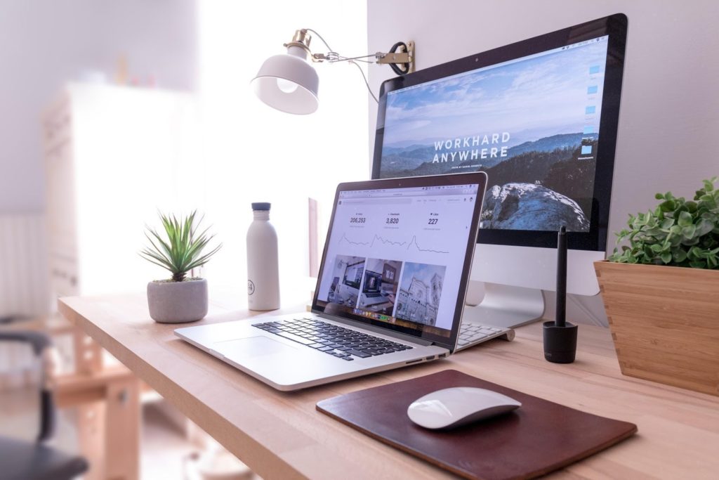 a laptop open on a desk with a larger PC screen turned on behind it