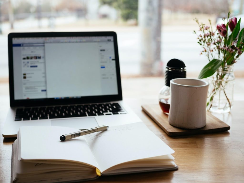 a laptop opened on a desk with a notebook, pen and coffee