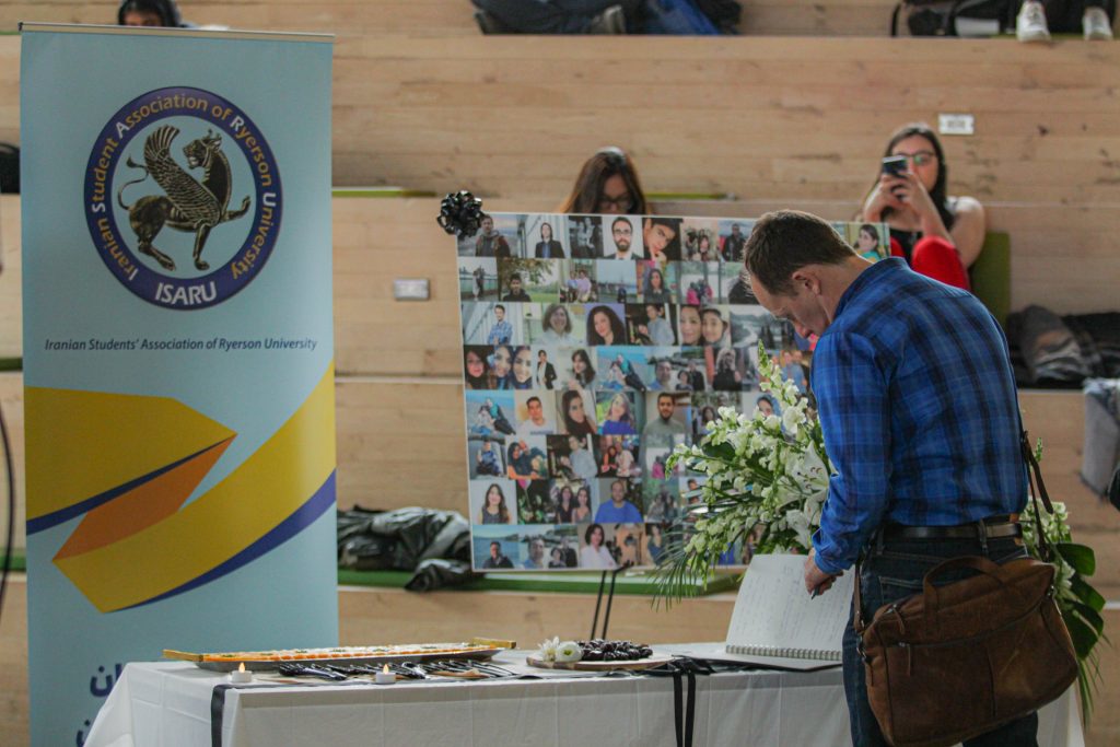 A man stands in front of a table with a sign reading Iranian Student Association of Ryerson University and a board with photos on it