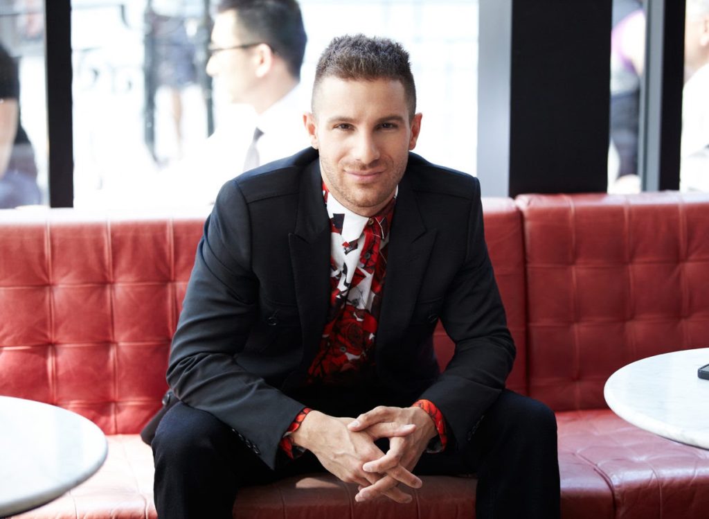 a man sits on a red couch smiling straight at the camera with his fingers interlocked