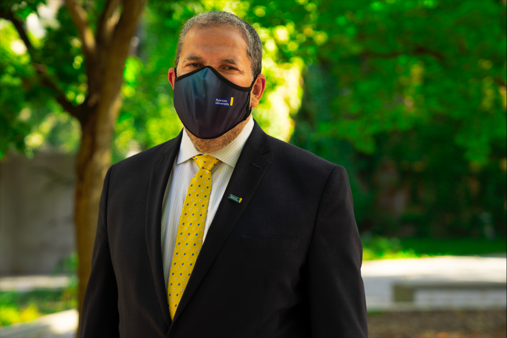 a man stands in front of a green background of trees on a sunny day wearing a black face mask