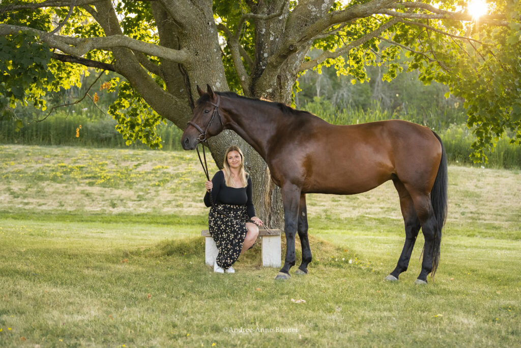 a blonde woman and her brown horse sit in front of a tree in a green open area on a sunny day