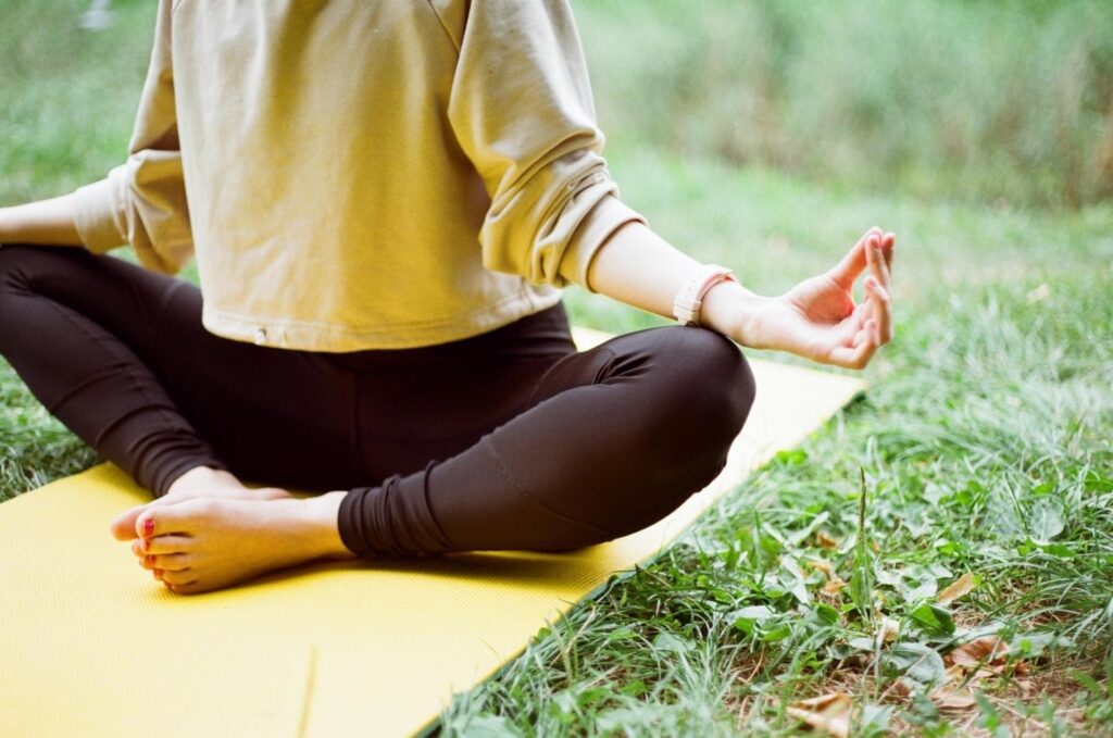 a person sits on a yoga mat laid out on the grass with their legs crossed and hands over their knees as though they are praying