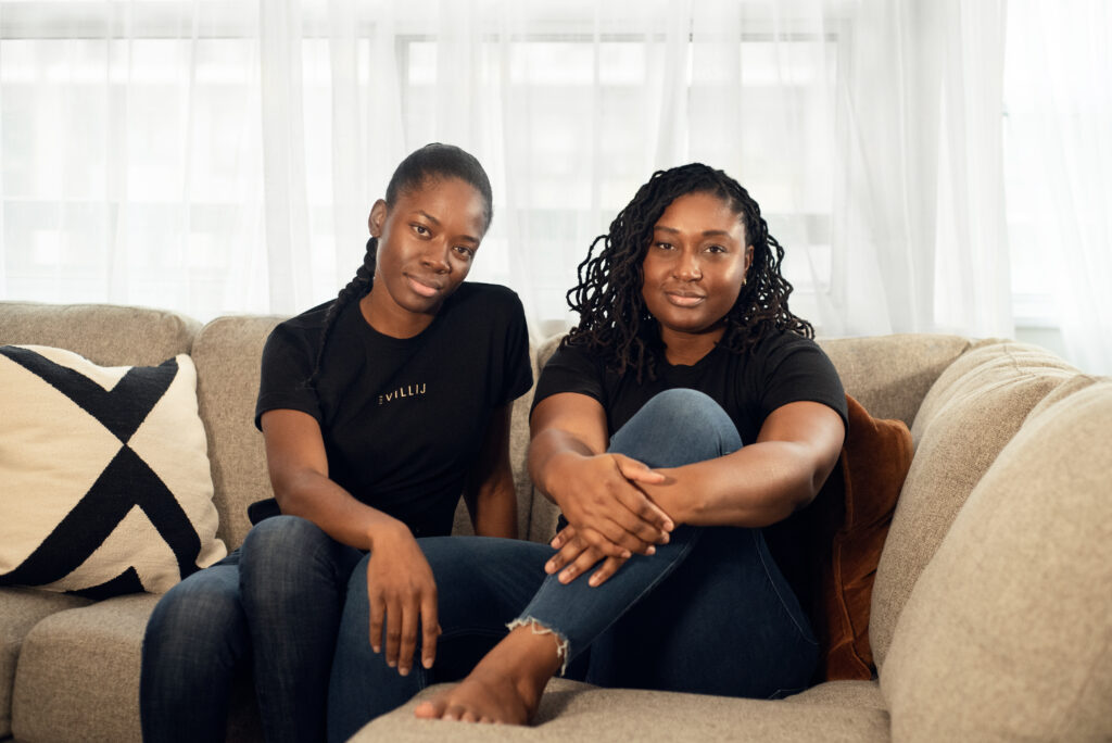 two women sit on a couch and look directly at camera